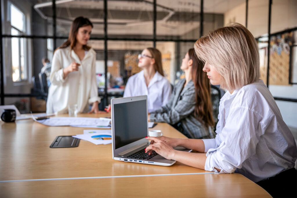 Grupo de mujeres jóvenes en una reunión de trabajo en una oficina moderna, una de ellas usando portátil mientras otras conversan