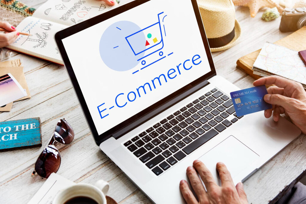 Ordenador portátil sobre una mesa de madera con la palabra 'E-Commerce' en pantalla y una ilustración de carrito de compra, mientras una persona sostiene una tarjeta de crédito, representando el servicio de creación de tiendas online WordPress de Ribsweb.