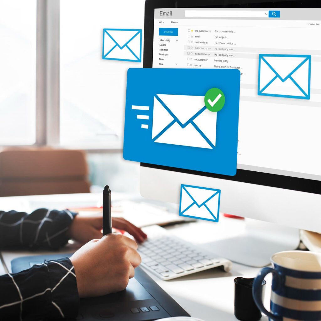 Persona utilizando una tableta gráfica frente a un ordenador con bandeja de entrada de correo electrónico abierta y sobres flotantes, simbolizando envío y gestión de emails