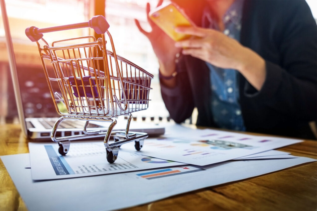 Carrito de la compra en miniatura sobre documentos con gráficos, junto a una persona usando un smartphone frente a un portátil, representando el comercio electrónico y la toma de decisiones digitales en ventas