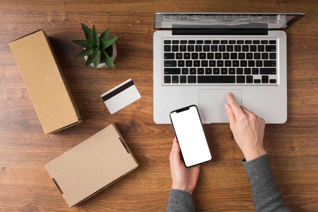 Persona usando portátil y smartphone para gestionar pedidos online, con cajas de envío, tarjeta bancaria y planta decorativa sobre mesa de madera, representando una tienda PrestaShop en funcionamiento.