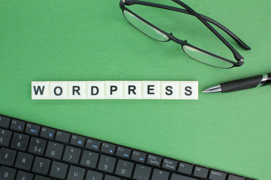 Palabra WordPress escrita con fichas sobre fondo verde, acompañada de teclado, bolígrafo y gafas: diseño web profesional con WordPress