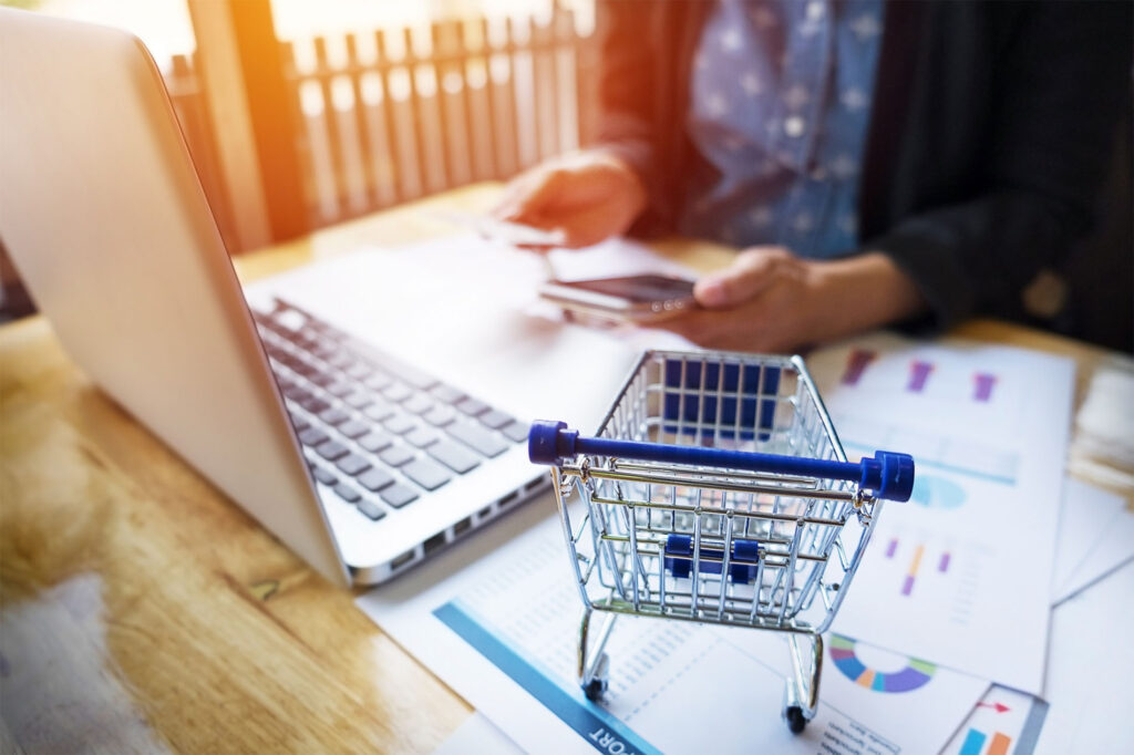 Escena de trabajo con portátil, gráficos de análisis y una persona sosteniendo una tarjeta de crédito, junto a un carrito de compras en miniatura sobre la mesa, simbolizando la gestión y optimización de tiendas online con PrestaShop.